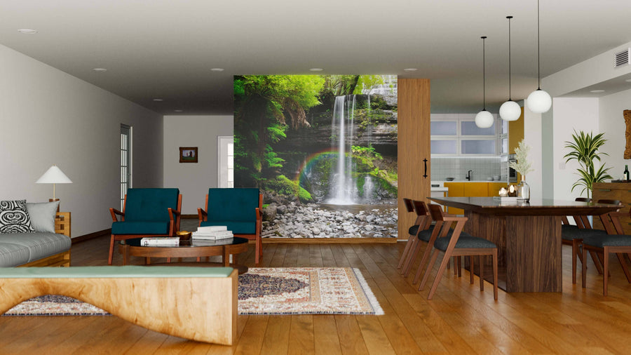 A modern open-plan living and dining room with wooden floors, blue chairs, a dining table, and a Waterfalls Wall Mural with lush greenery and a rainbow creates a stunning nature-inspired focal point in the space.
