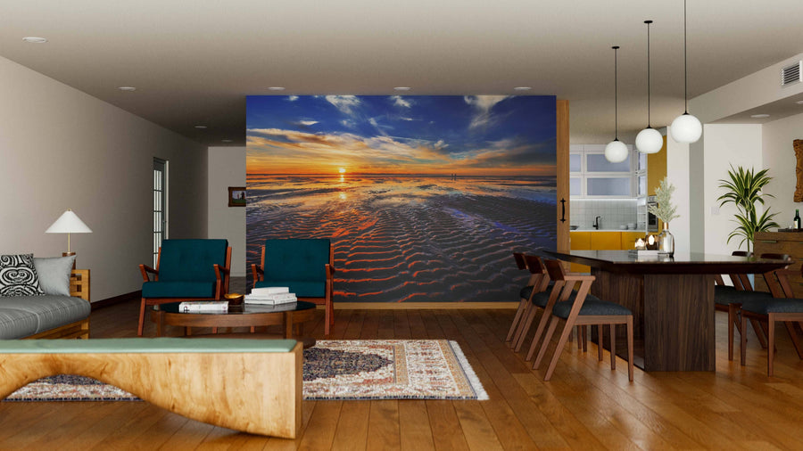 A modern living-dining room features wooden floors, stylish furniture, warm lighting, and a large Waves on the Sand Wall Mural displaying a vivid sunset over rippled sand and ocean waves, creating a relaxing atmosphere.