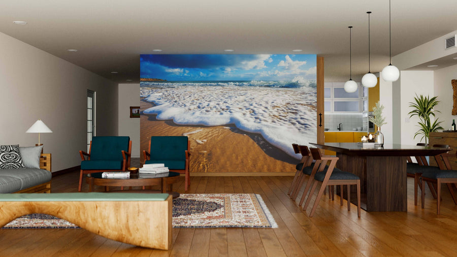 A modern open-plan living and dining room with wooden floors, teal armchairs, a large dining table, and the vibrant Wet Feet Wall Mural featuring sandy beach and ocean waves, creating a lively coastal atmosphere.