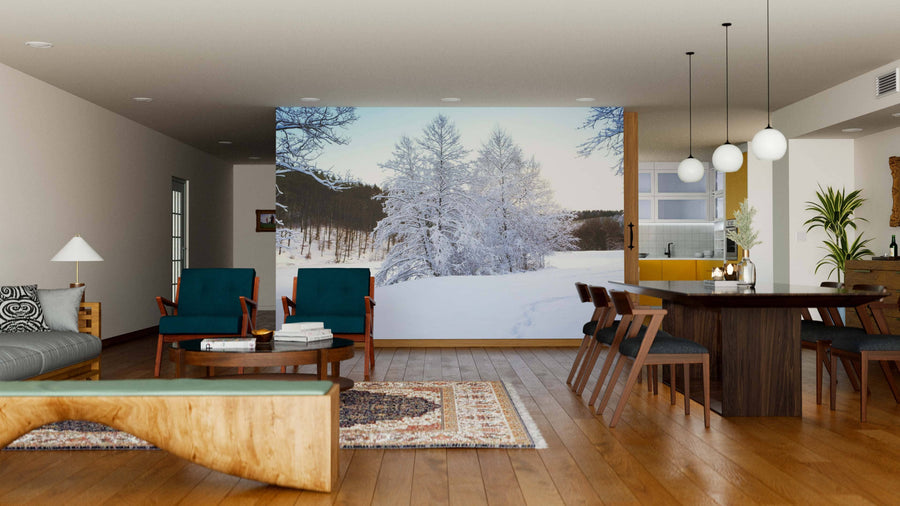 A modern open-plan living and dining room with wooden floors features teal chairs, a gray sofa, and round pendant lights. The focal point is the "Winter Landscape with Snow-Covered Trees Wall Mural," adding cozy, wintry charm.