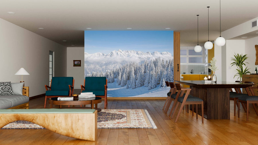 A cozy, modern living room with green and brown furniture features the "Winter in the Alps Wall Mural." Large windows show snowy pines and mountains under a blue sky, while warm lighting contrasts with the wintry scene outside.