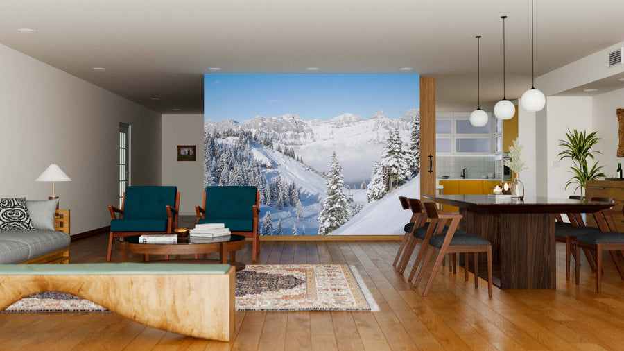A modern living and dining area with wood floors features blue chairs, a wooden bench, a dining table under pendant lights, and the “Winter in the Swiss Alps Wall Mural,” showcasing snowy mountains beneath a clear blue sky.