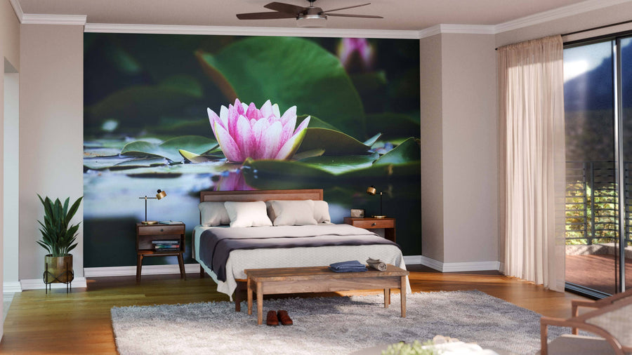 A cozy bedroom with wood furniture, cream walls, and a large Lotus Flower Wall Mural of pink lotus on lily pads behind the bed. Sunlight filters through sheer curtains over sliding doors, while a plant, rug, and bench add warmth to the serene space.