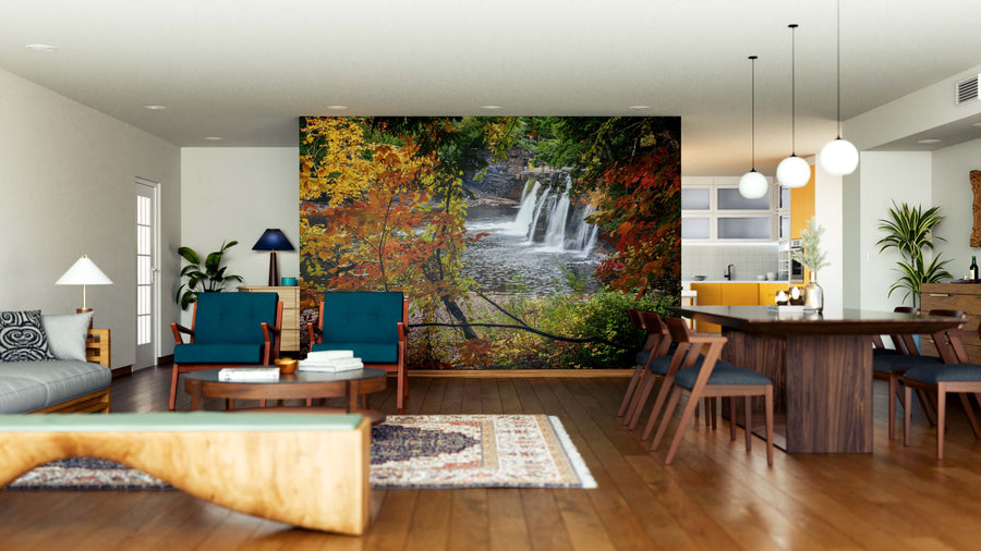 A modern living-dining room with wooden floors features the Manabezho Falls Wall Mural. Mid-century teal furniture, a wooden coffee table, indoor plants, and pendant lights create a cozy and stylish atmosphere.