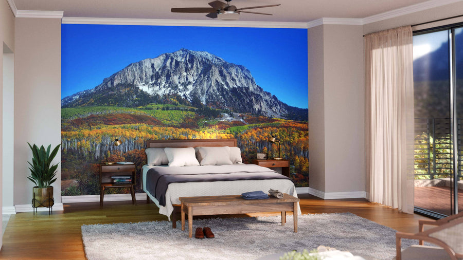 A cozy bedroom showcases the Marcelina Mountain Over Aspen Forest Wall Mural behind the bed, with neutral walls, wooden furniture, a potted plant, and sheer curtains on the glass door creating a warm, inviting atmosphere.