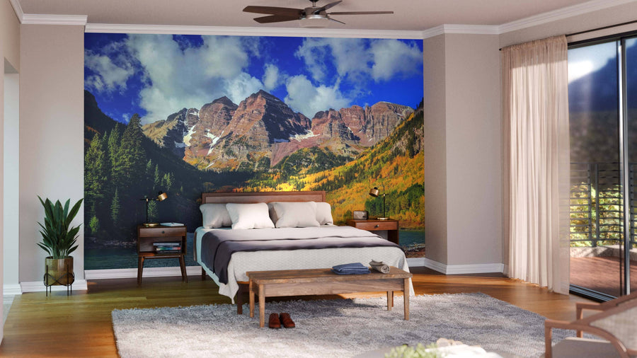 A spacious bedroom features a Maroon Bells in Colorado Wall Mural behind the bed, complemented by a bench, plant, lamps, soft rug, wood floor, ceiling fan, and glass doors with beige curtains for an inviting atmosphere.