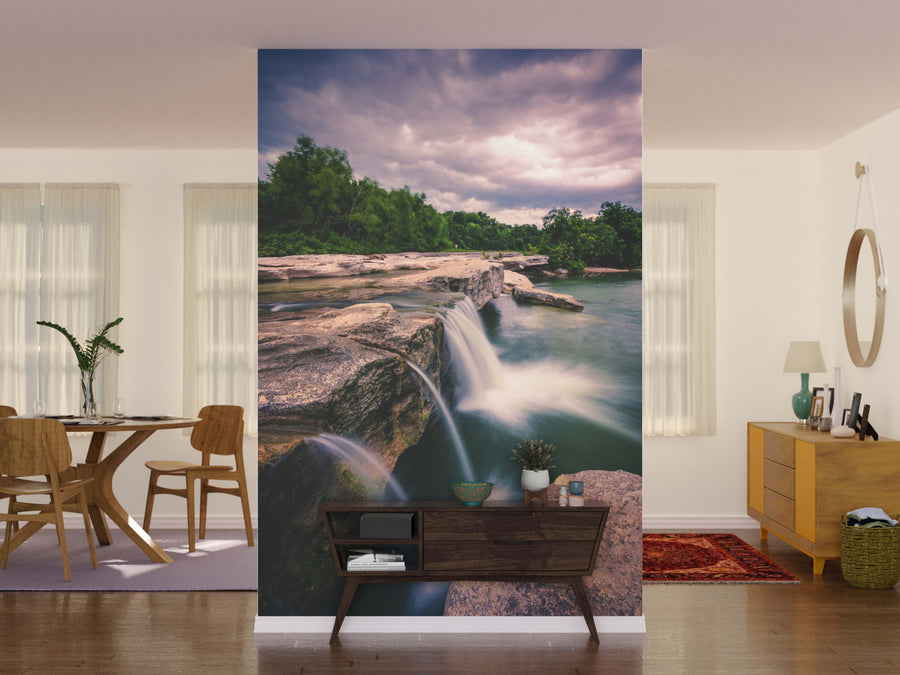 A modern living and dining room showcases the McKinney Falls Wall Mural, featuring a lush green river and waterfall. Wooden furniture, plants on a small table, soft lighting, a round mirror, and a vibrant rug complete the inviting space.