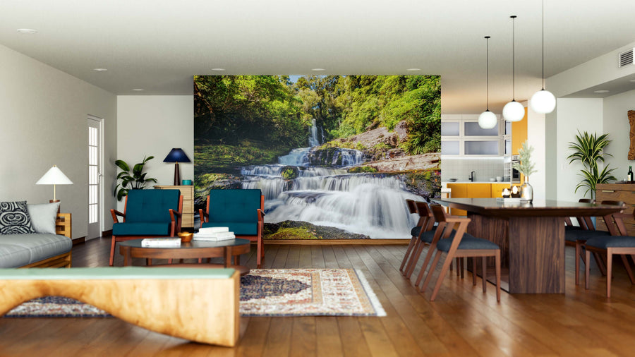 A modern living and dining area with wooden floors, blue and wood furniture, a patterned rug, pendant lights, and plants is highlighted by the Mclean Falls Wall Mural—a lush forest waterfall as the natural focal point.