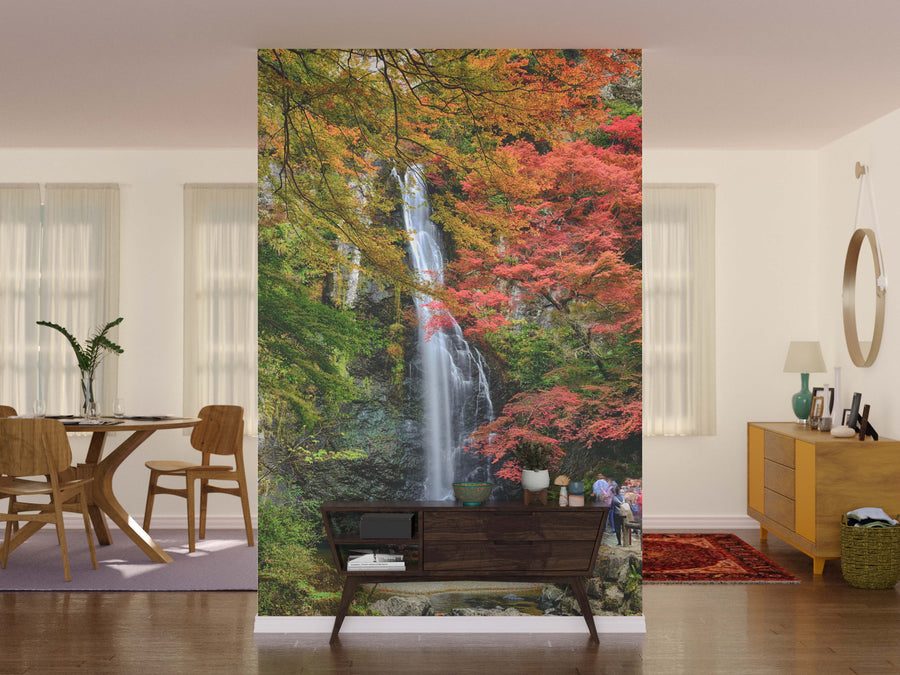 A living room with neutral walls and natural light features the vibrant Minoo Waterfall Wall Mural framed by red, orange, and yellow foliage. A modern wood console sits in front, with a dining table to the left and dresser to the right.