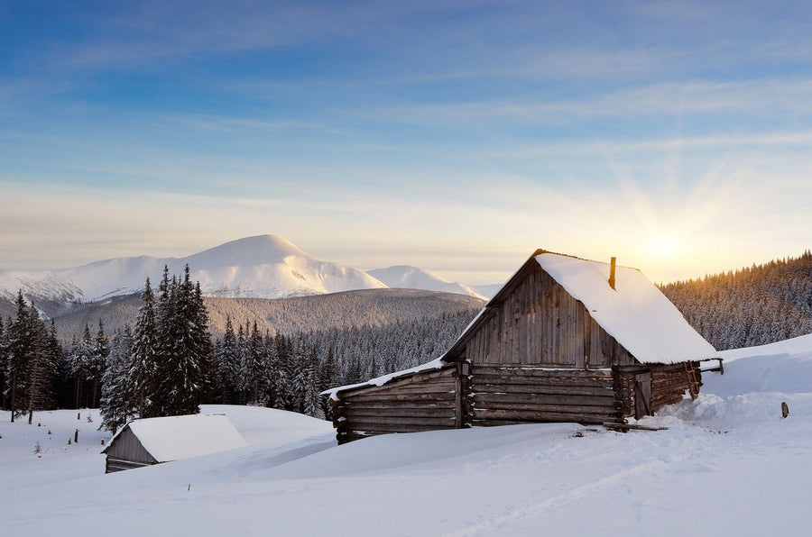 The "Cabin in the Mountains Wall Mural" showcases a peaceful winter scene with a snow-covered cabin and evergreen trees, set against majestic sunlit mountains under a clear blue sky, ideal for custom printed wall murals.