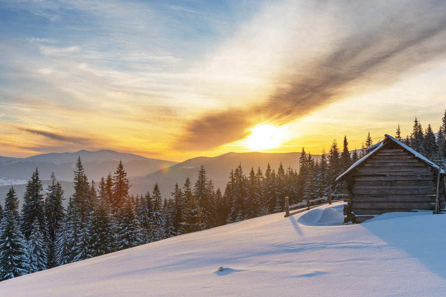 A rustic wooden cabin in a snowy forest is illuminated by sunrise, as dramatic clouds streak across the sky. Distant mountains complete this serene winter view, reminiscent of the "Severe Winter on a Mountain Range Wall Mural.