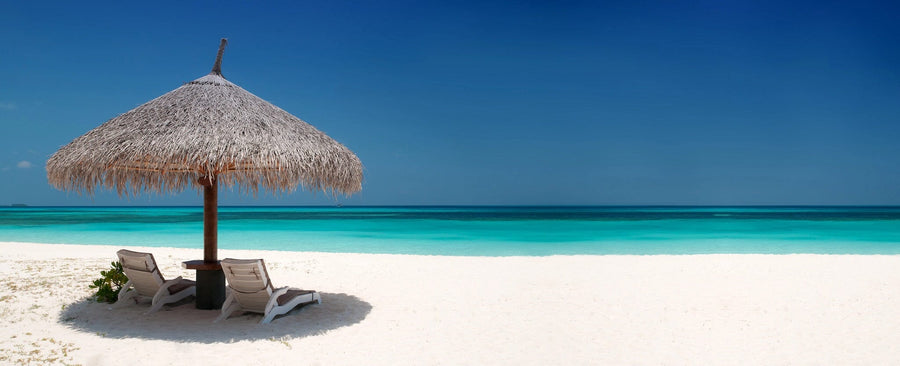 The Beach Chairs and Umbrella Wall Mural depicts two lounge chairs under a thatched umbrella on a pristine white sandy beach, facing the turquoise ocean. With a clear blue sky overhead and a small table between the chairs, it evokes a serene and tropical getaway.