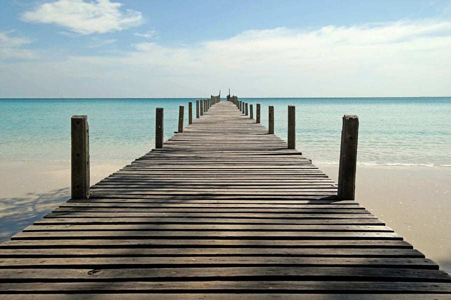 A partly cloudy sky arches over a wooden pier reaching into a turquoise ocean, flanked by weathered posts. This serene beach scene, with gentle waves kissing the shore, is reminiscent of the Beach Pier Wall Mural, an ideal peel and stick wallpaper design for tranquility.