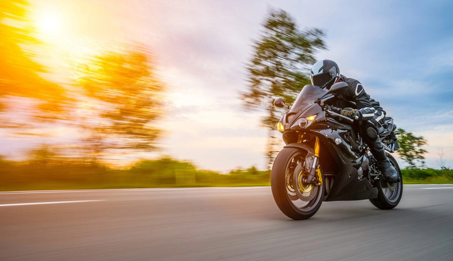 A motorcyclist in black gear speeds on a sleek, black sport motorcycle down a paved road. The blurred trees and vivid sunset evoke the motion of a Motor 18 Wall Mural, with partly cloudy skies in warm hues enhancing the dynamic scene.