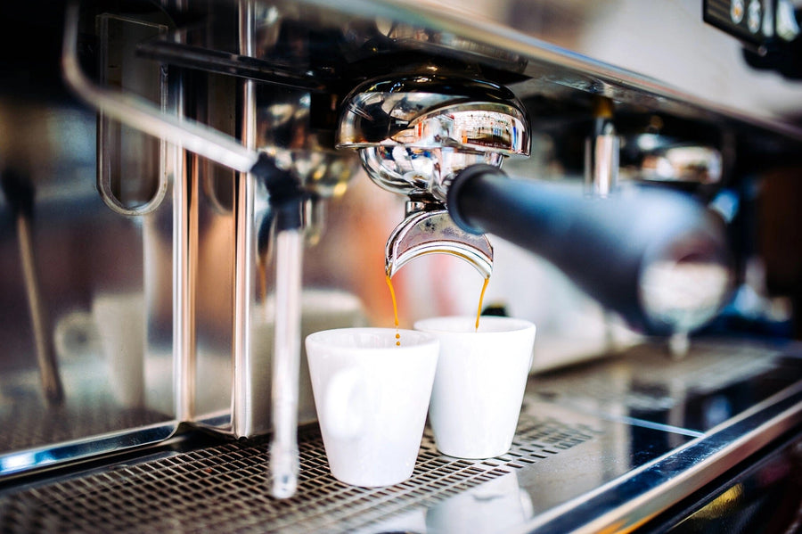 A close-up of an espresso machine brewing coffee into two white cups against a backdrop reminiscent of the Coffee 10 Wall Mural. The metallic spout drips rich espresso, with a black handle extending towards the viewer, as light highlights the shiny surface and drip tray below.