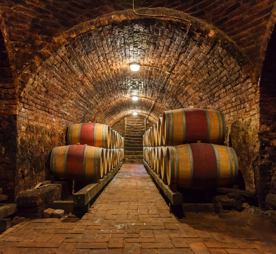 A dimly lit wine cellar with a vaulted brick ceiling boasts rows of wooden barrels with metal hoops along a brick-paved path. Soft, warm lighting enhances the textured bricks and wood, while the Brick-Walled Wine Cellar Wall Mural adds charm to this cozy atmosphere.