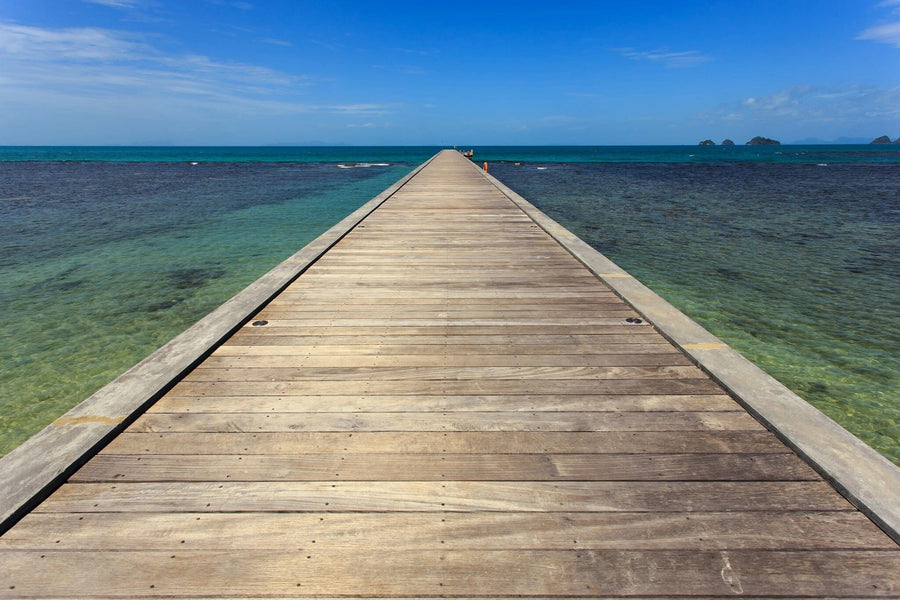 A wooden pier extends into the clear ocean, resembling the "Bridge to the Sea Wall Mural" by Deposit Photo. The water blends turquoise and deep blue with small islands in sight, giving an endless perspective.