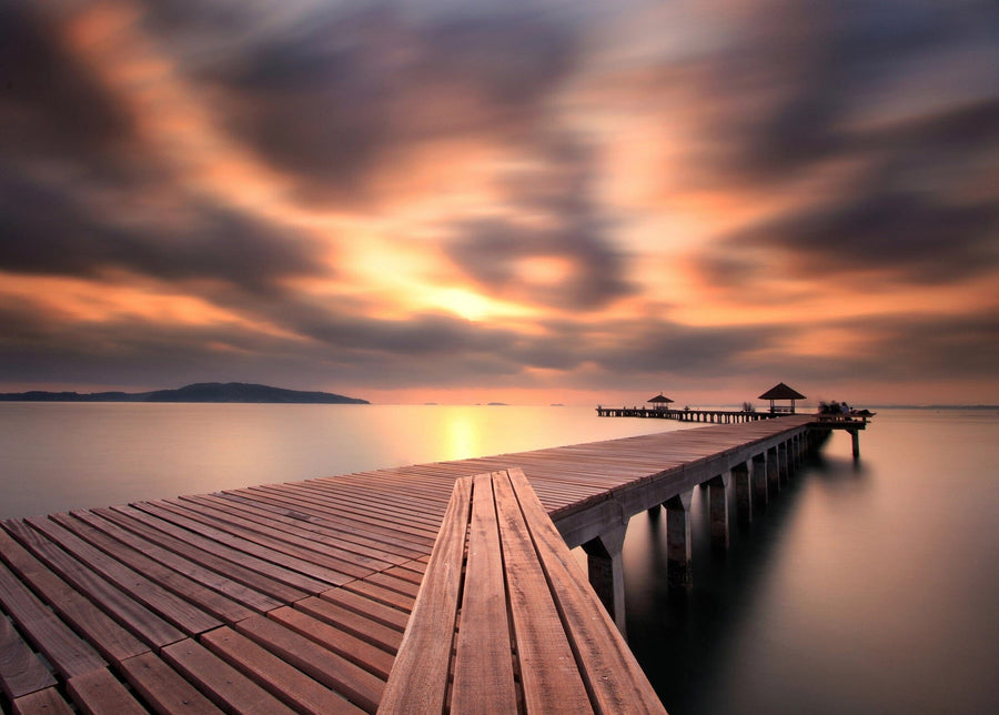 The Calm Ocean Walkway Wall Mural features a long pier stretching over tranquil waters towards gazebos, beneath a vibrant sunset sky. Clouds resemble dynamic patterns of peel and stick murals, with a faint land silhouette on the horizon adding to the serene and picturesque scene.