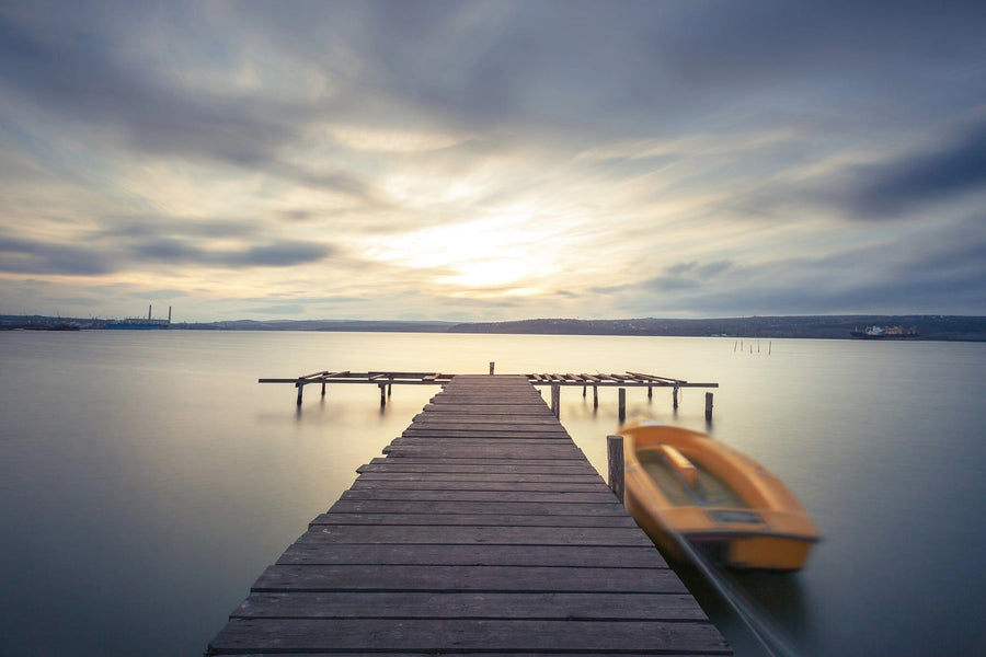 The "Calm Waters at Sunset Wall Mural" by Deposit Photo showcases a tranquil landscape with a wooden pier extending into a calm lake under a cloudy sky. A blurred orange boat adds dynamic movement to the serene scene as the setting sun casts a soft glow on the horizon.