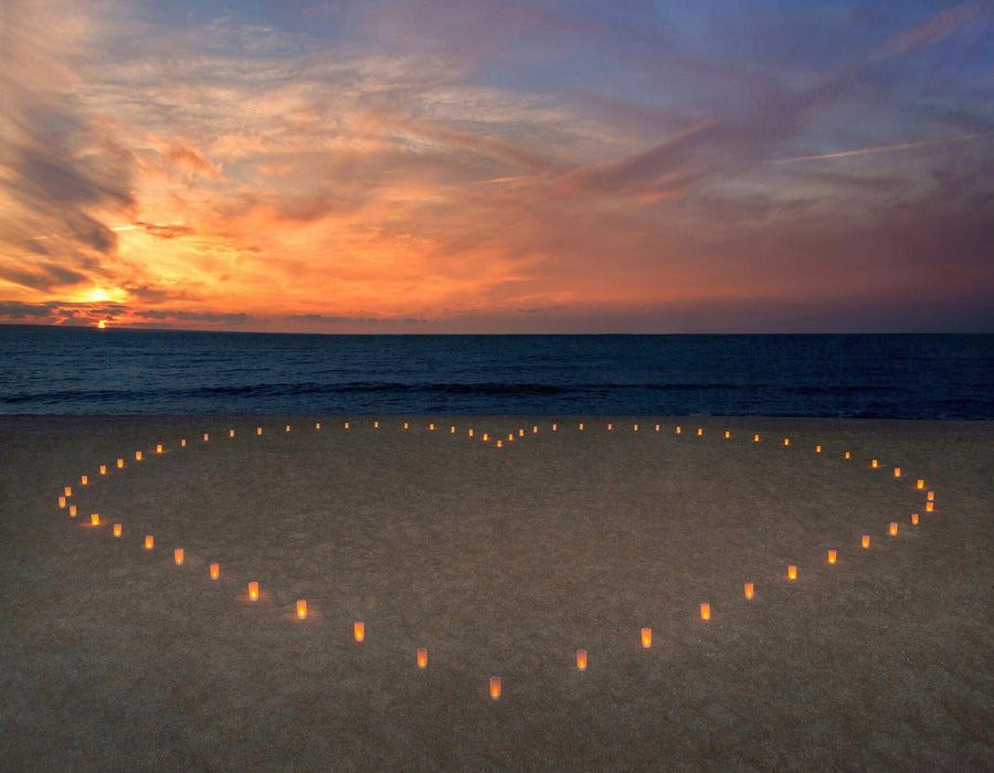 The Candles Heart Wall Mural depicts lit candles forming a heart on a sandy beach at sunset, with the orange and pink hues of the sky mirrored in the tranquil ocean, creating a romantic and peaceful atmosphere.