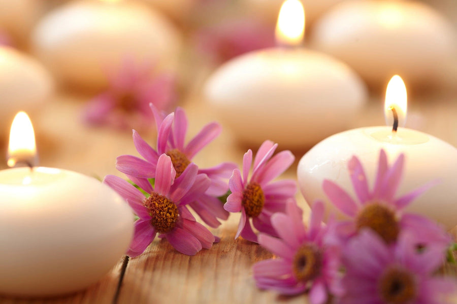 The Candles and Flowers Wall Mural depicts a softly lit scene with round candles glowing warmly on a wooden surface, surrounded by pink daisies. The blurred background enhances the serene ambiance, highlighting the gentle colors and textures of flowers and candlelight.