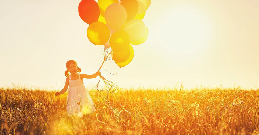 A young girl in a white dress with yellow and orange balloons stands in a sunlit field of tall grass, evoking the dreamy essence of the "Inspirational 2 Wall Mural," as her joyful smile glows warmly in the sunset's embrace, highlighting the summer atmosphere.