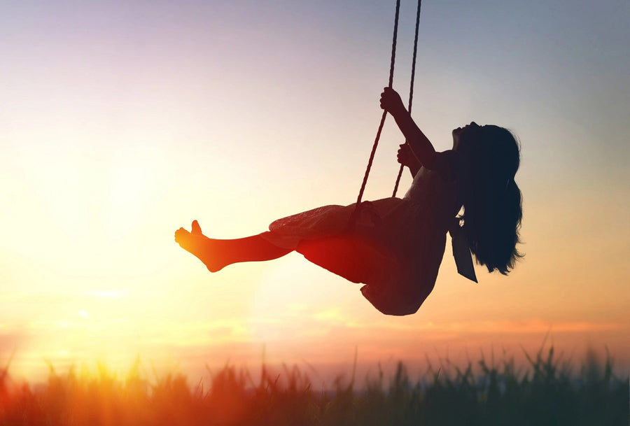 The "Child on Swing Wall Mural" features a joyful child in a dress with a bow on a swing, silhouetted against an orange, pink, and purple sunset. The peel and stick wallpaper depicts the sun low on the horizon with tall grass below.