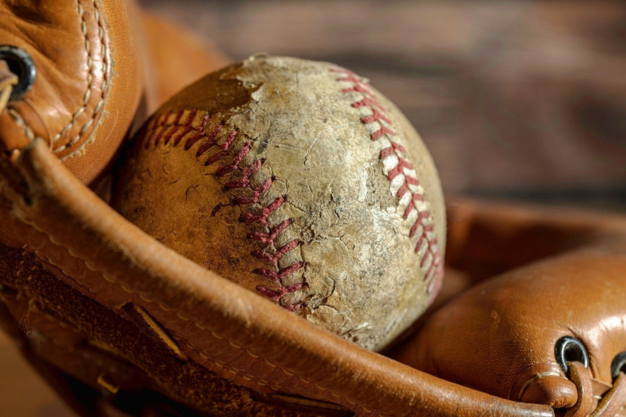 The Classic Leather and Stitch Wall Mural features a close-up of a weathered baseball with red stitching in a well-used brown glove, creating nostalgic charm for any room with its soft, blurred background. Peel and stick for easy decor.