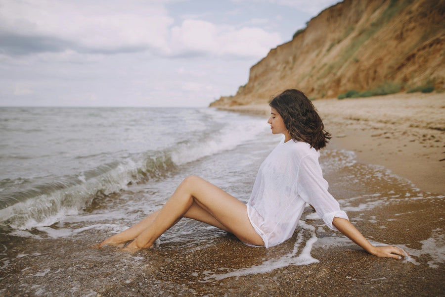 A woman in a sheer white shirt sits on wet sand as waves touch her legs beneath cliffs and a cloudy sky. Bring this tranquil seaside scene home with the Coastal Serenity Wall Mural for a calming, custom-printed feature wall.