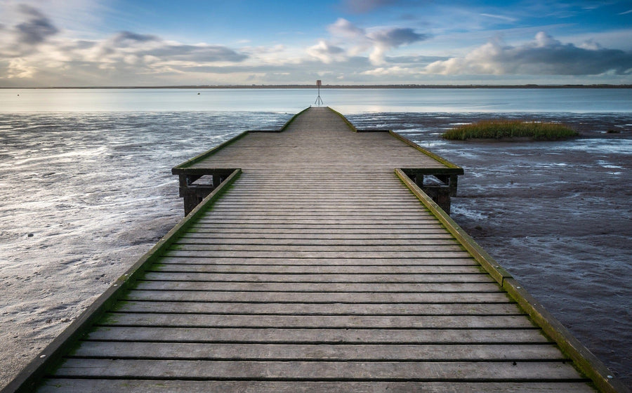 A long wooden pier with green edges stretches over a muddy shore toward calm blue water beneath a partly cloudy sky, with grass on the left and a distant horizon—featured in our Coastal Solitude Wall Mural for peaceful spaces.