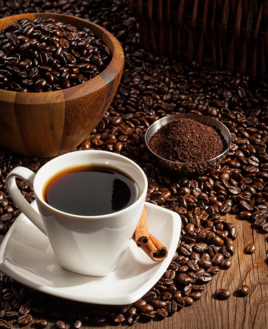 A Coffee Cup with Cinnamon Stick Wall Mural features a steaming black coffee on a white saucer, surrounded by coffee beans and ground coffee in bowls, with a cozy café scene depicted on the rustic wooden surface.