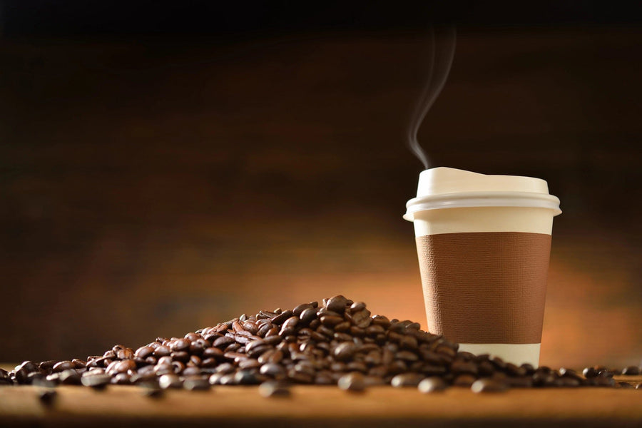 A steaming takeaway coffee cup with a brown sleeve sits next to scattered beans on wood, set against a warm, blurred gradient backdrop. The scene evokes a cozy atmosphere similar to those featured in the Coffee 13 Wall Mural