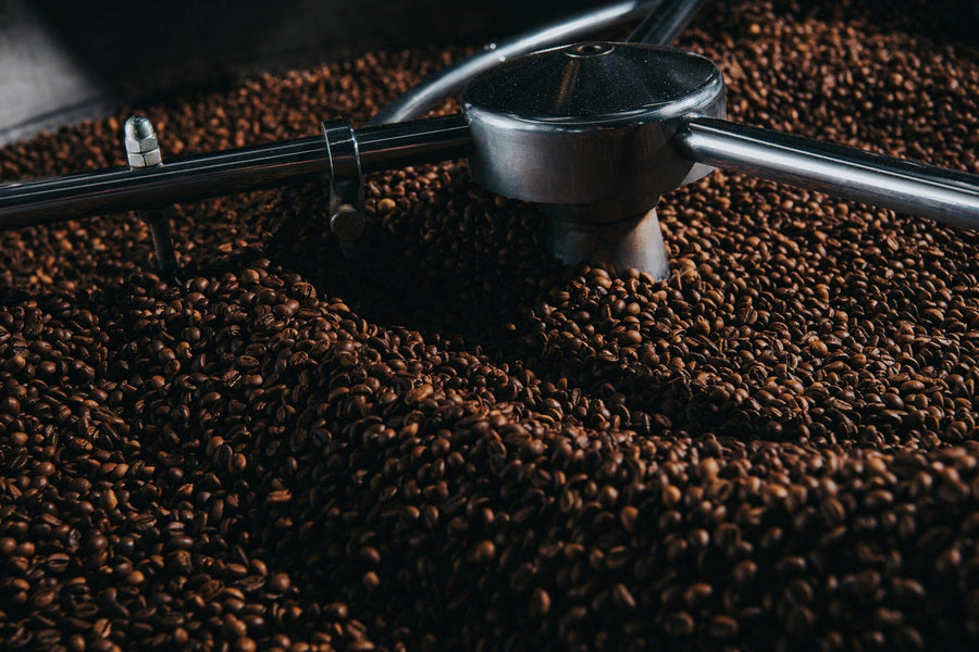 A large metal roaster brimming with dark roasted coffee beans, stirred by a rotating arm. Dim lighting enhances the warm ambiance, highlighting the beans' glossy texture against a backdrop featuring the Coffee 8 Wall Mural.