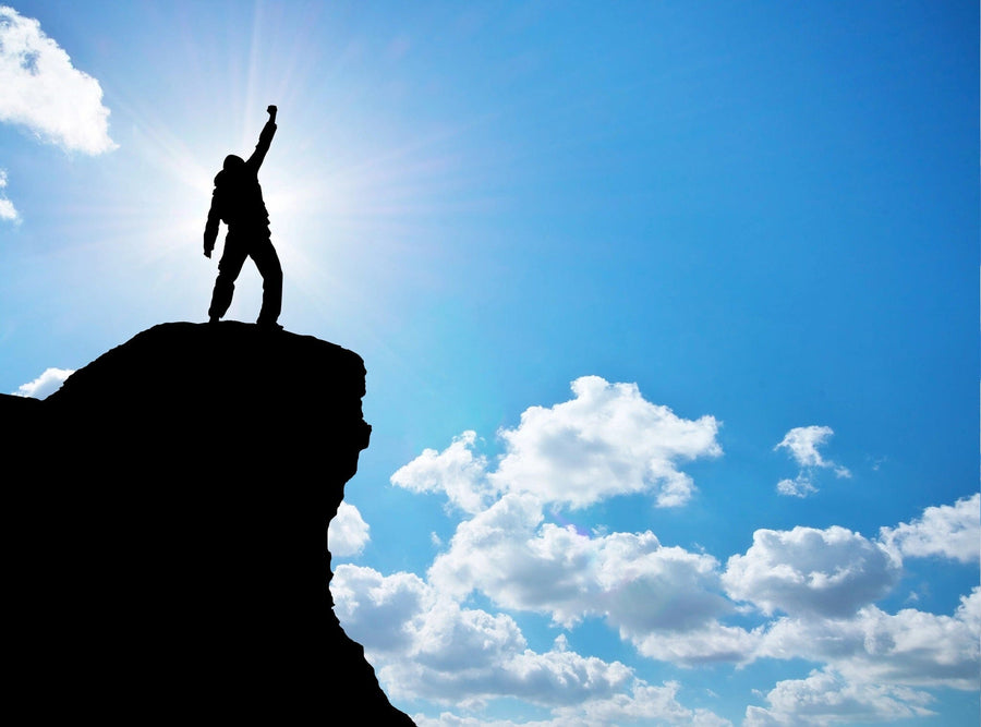 A person stands triumphantly on a rocky cliff against a clear sky, reminiscent of the breathtaking scene from the Inspirational 4 Wall Mural. The sun shines brightly behind them with scattered clouds, and one arm raised in victory symbolizes achievement and success.