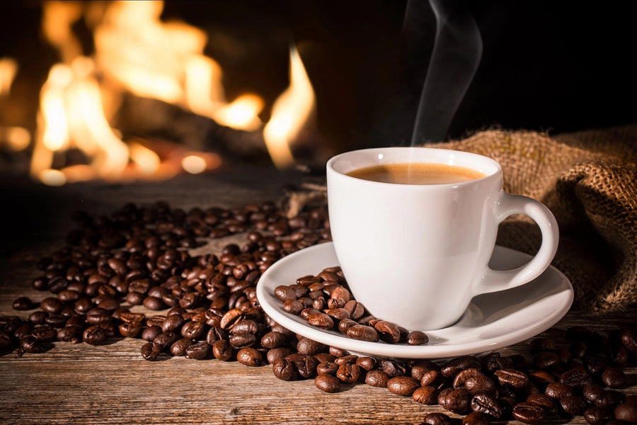 A steaming white coffee cup rests on a saucer amid scattered beans on a rustic wooden table adorned with  Cozy Coffee Wall Mural. The blurred flames of a cozy fireplace create a warm ambiance, while a burlap sack is partially visible on the left.