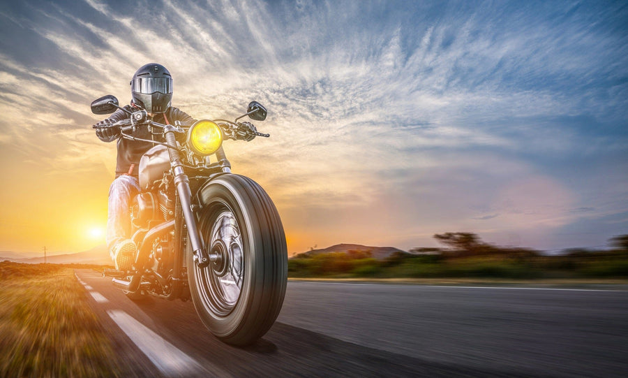 A motorcyclist wearing a helmet rides a cruiser motorcycle on an open road under the sun's warm glow and dramatic cloud patterns, resembling the "Cruising into Sunset Wall Mural," with the headlight on as they lean right, chasing the endless horizon.