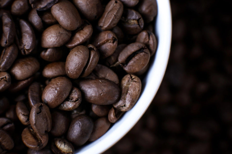 Close-up of glossy dark brown coffee beans in a white bowl, set in the upper right corner with blurred beans behind. Ideal for decorating your space with the Cup of Coffee Beans Wall Mural—available as custom prints or peel and stick murals.