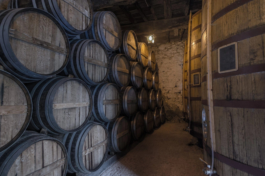 In a dimly lit cellar, with stone walls adding rustic charm, rows of stacked wooden barrels line the right, and a large wooden tank sits on the left. A custom Wine 19 Wall Mural enhances the space as shadows from a single bulb amplify the ambiance.