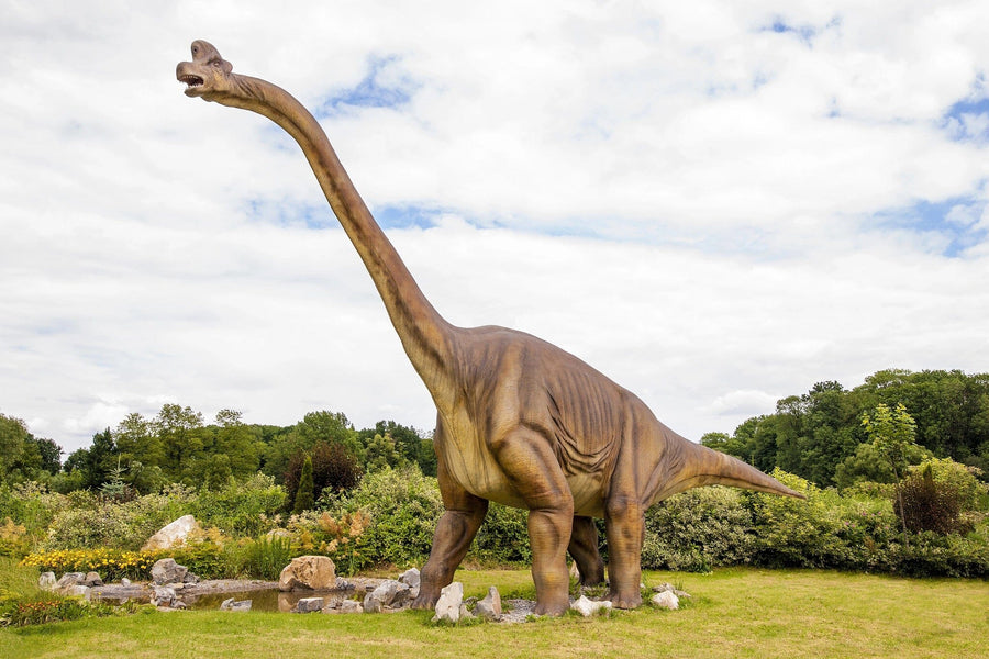 A large, realistic dinosaur statue with a long neck and four legs stands on grassy terrain among bushes and rocks under partly cloudy skies. Captured in profile, this scene feels like stepping into the "Dinosaur Sculpture Wall Mural", where trees whisper tales of the past.