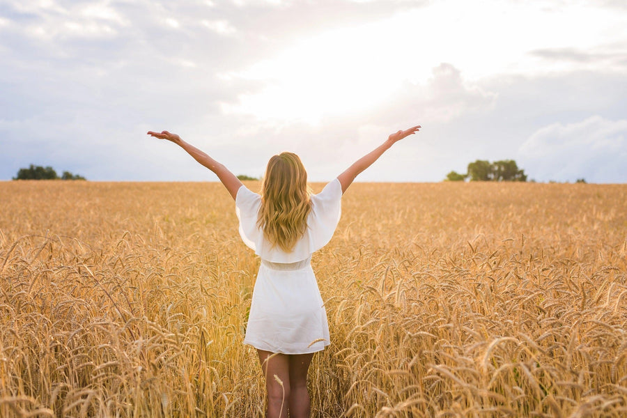 A woman in a white dress with arms outstretched stands in a golden wheat field under a luminous sky, embodying the serenity of the Inspirational 4 Wall Mural. Her back is to the camera, facing bright clouds and distant trees as the sun peeks through.