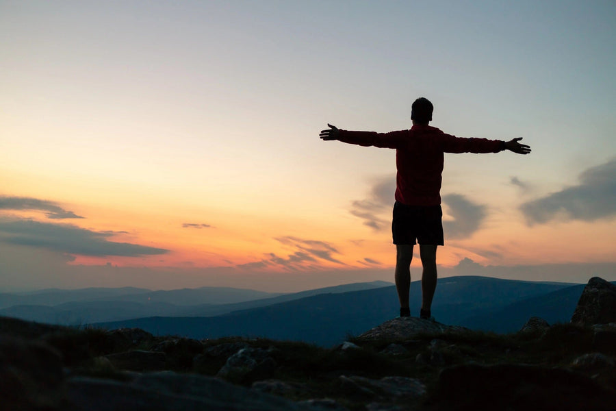 A person stands on a rocky hill, arms open to a vibrant sunset, reminiscent of the Inspirational 6 Wall Mural. The sky shines in orange, pink, and blue hues against distant mountains. They're dressed in shorts and a long-sleeved shirt.