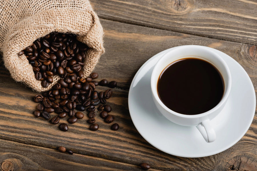 The Coffee 6 Wall Mural features a white cup of black coffee on a saucer, with dark roasted beans spilling from a burlap sack—ideal for adding fresh coffee vibes to your space with this custom printed wallpaper.