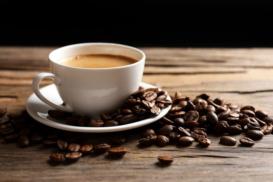 A white ceramic cup of coffee sits on a matching saucer on a wooden table, surrounded by scattered coffee beans. This rustic scene echoes the charm of the "Coffee 6 Wall Mural". The dark background enhances its contrast and warmth.