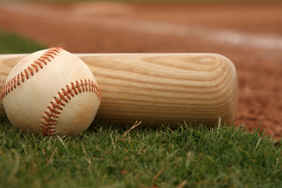 A close-up of a wooden baseball bat and a white ball with red stitching on green grass, set against a blurred stadium infield—Game Day Ready Wall Mural, ideal for adding sporty flair to your space with easy peel-and-stick application.