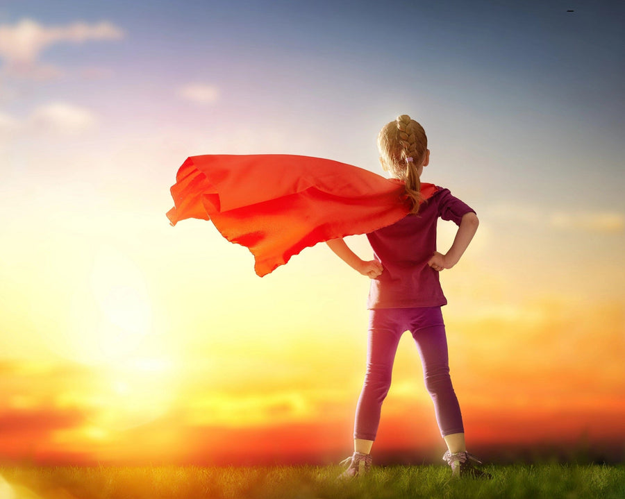 A child confidently stands on grass facing a vibrant sunset, akin to art on "Girl Plays Superhero" wall mural. Wearing a red cape flowing in the breeze and hands on hips, the orange and blue sky enhances the heroic mood, symbolizing strength and imagination.