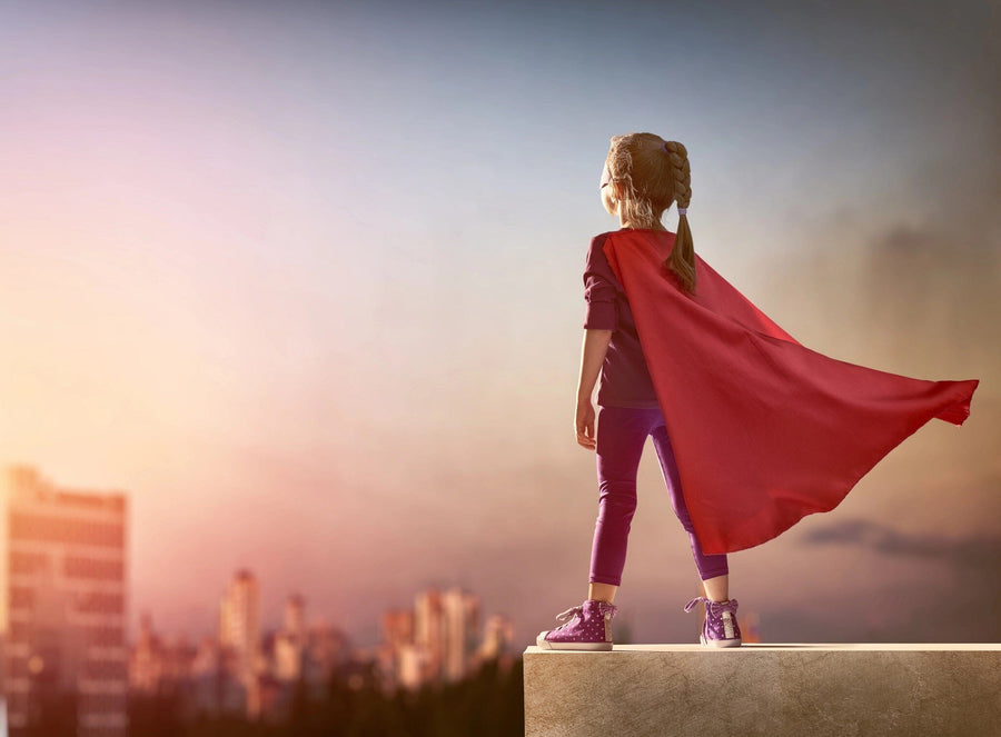 A young girl stands on a ledge, her red cape fluttering against a sunset city skyline. In purple leggings and sparkly sneakers, she captures the spirit of adventure—an image as stunning as the "Girl Superhero Wall Mural"