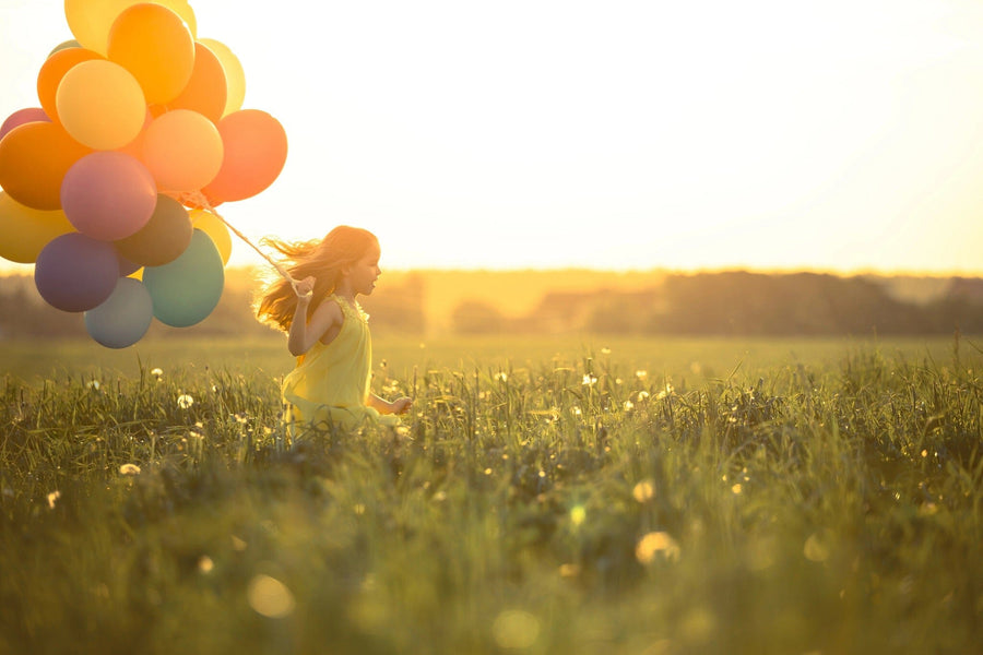 A young girl in a yellow dress joyfully runs through a sunlit grassy field with balloons, embodying the serenity of Happy Girls Wall Mural, where warm sunlight and distant trees under a clear sky enhance the scene.