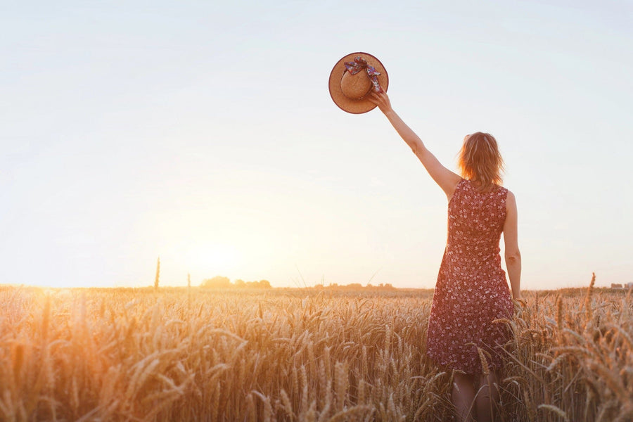 A woman in a sleeveless floral dress stands in a golden wheat field at sunset, raising a ribboned straw hat skyward as she faces away, embodying the tranquility captured in our Golden Fields Wall Mural to bring serene beauty into any space.