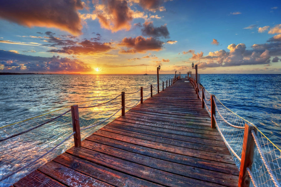 The Pier 1 Wall Mural depicts a serene wooden pier stretching into a vast ocean during a vibrant sunset, with orange and pink hues gracing the partially cloudy sky. Rope railings add to the tranquil scene as the sun nears the horizon, casting calming reflections on the water.