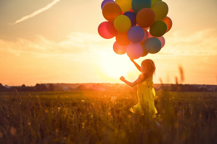 A young girl in a yellow dress holds colorful balloons in a field at sunset, with the sky's orange and pink hues resembling the "Happy Girl Wall Mural" The vibrant balloons beautifully contrast against the serene backdrop.